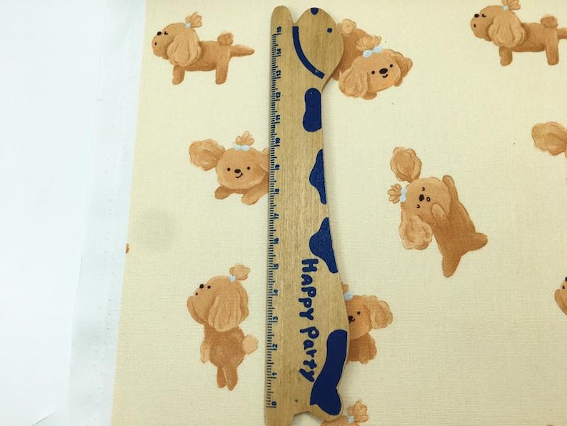 May include: A light beige fabric with a repeating pattern of cartoon dogs in various poses. A wooden ruler with blue accents and the text "Happy Party" is placed vertically on the fabric.