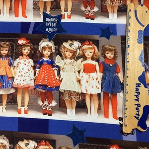Op de afbeelding: Een kleurrijke stof met een herhalend patroon van poppen die jurken en hoeden dragen. De poppen staan op een blauw-wit geruite achtergrond. De stof heeft de tekst "Under Milk Wood" en "Happy Party" erop gedrukt.