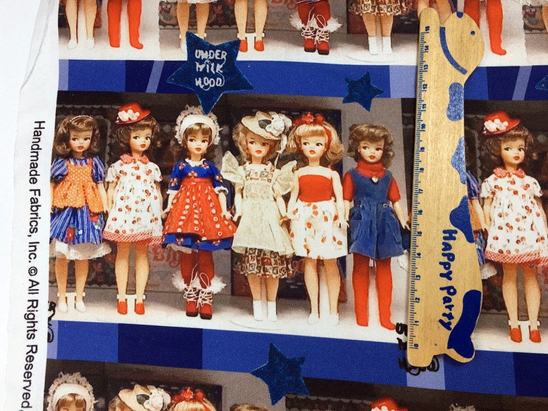 Op de afbeelding: Een kleurrijke stof met een herhalend patroon van poppen die jurken en hoeden dragen. De poppen staan op een blauw-wit geruite achtergrond. De stof heeft de tekst "Under Milk Wood" en "Happy Party" erop gedrukt.