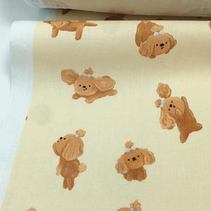 May include: Off-white fabric featuring a repeating pattern of cartoon-style brown dogs with blue bows. The dogs are in various poses, some standing, some lying down, and some with their paws raised. The fabric is folded over.