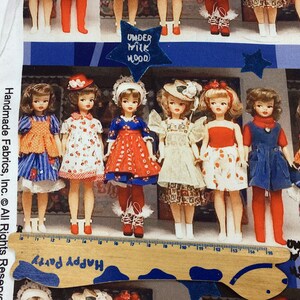 Op de afbeelding: Een stoffen paneel met een rij vintage poppen gekleed in kleurrijke jurken met rode, witte en blauwe accenten. De poppen staan voor een blauw-wit gestreepte achtergrond met de tekst "Under Milk Wood" erop gedrukt.