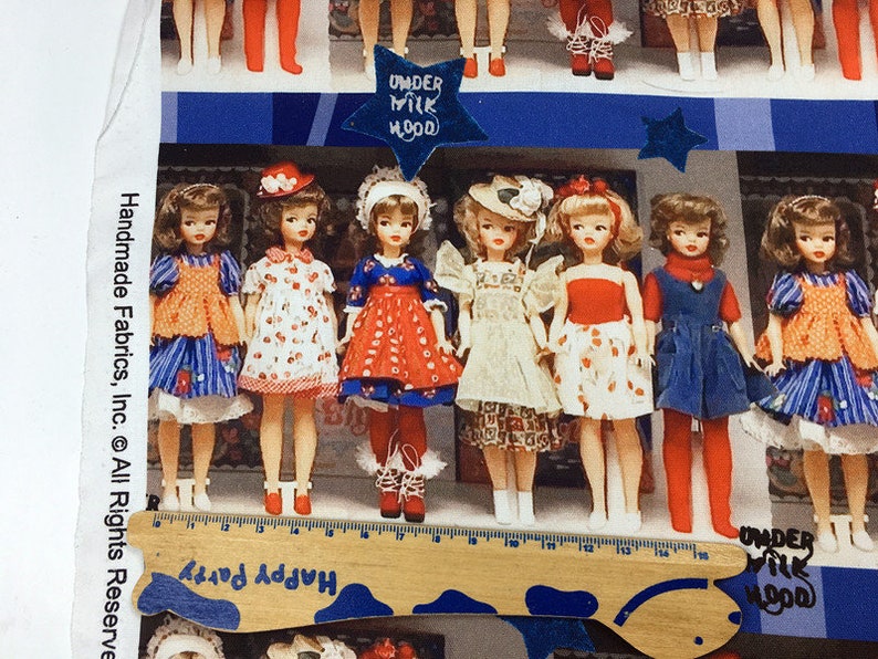Op de afbeelding: Een stoffen paneel met een rij vintage poppen gekleed in kleurrijke jurken met rode, witte en blauwe accenten. De poppen staan voor een blauw-wit gestreepte achtergrond met de tekst "Under Milk Wood" erop gedrukt.