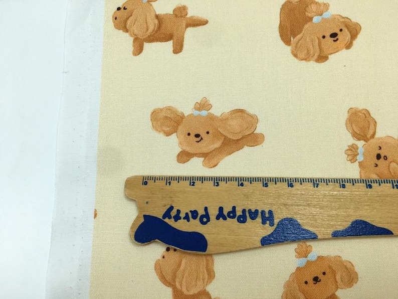 May include: Fabric featuring a repeating pattern of cartoon poodle dogs in shades of brown, with blue bows. A wooden ruler with the text "Happy Party" is placed on the fabric. The fabric has a cream-colored background.
