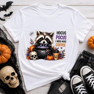 Può includere: Una t-shirt bianca con la grafica di un procione con papillon, seduto dietro un calderone nero pieno di liquido viola. Il design include un teschio, zucche, fiori e il testo HOCUS POCUS I NEED MORE COFFEE.
