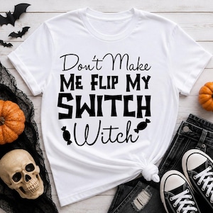 Op de afbeelding: Een wit T-shirt met korte mouwen en de zwarte tekst "Don't Make Me Flip My SWITCH Witch" met twee kleine zwarte snoepillustraties. Het shirt heeft een ronde hals en een standaard pasvorm met een grafisch ontwerp op de voorzijde.