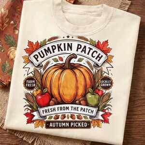 Könnte beinhalten: Ein cremefarbenes T-Shirt mit einer Grafik eines großen orangefarbenen Kürbisses, umgeben von roten und grünen Äpfeln, Maiskolben und Herbstblättern. Der Text lautet PUMPKIN PATCH, FARM FRESH, LOCALLY GROWN, FRESH FROM THE PATCH und AUTUMN PICKED.
