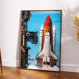 May include: A framed print of a space shuttle launching into a blue sky. The shuttle is white with orange and red accents, positioned on a launch pad with smoke and flames. The print is in a brown wooden frame.