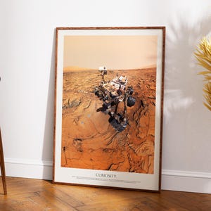 Könnte beinhalten: Ein gerahmter Druck mit dem Curiosity-Rover auf dem Mars. Das Bild zeigt den Rover in einer rotbraunen Landschaft mit dem Text "Curiosity" am unteren Rand. Der Druck befindet sich in einem braunen Holzrahmen und lehnt an einer weißen Wand.