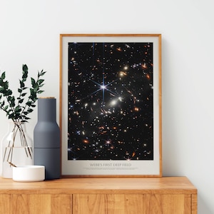 a framed poster of the Hubble Deep Field, a collection of galaxies captured by the Hubble Space Telescope. The poster displays a stunning view of the universe, showcasing the vastness and complexity of the cosmos. The poster is placed on a wooden shelf, accompanied by a vase of greenery and a white bowl, creating a harmonious and visually appealing display.