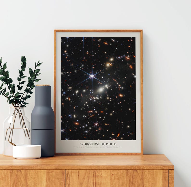 a framed poster of the Hubble Deep Field, a collection of galaxies captured by the Hubble Space Telescope. The poster displays a stunning view of the universe, showcasing the vastness and complexity of the cosmos. The poster is placed on a wooden shelf, accompanied by a vase of greenery and a white bowl, creating a harmonious and visually appealing display.
