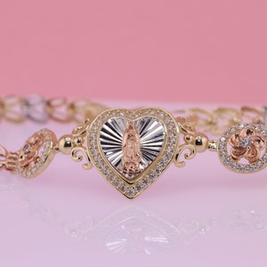 May include: A gold bracelet with a heart-shaped charm featuring a depiction of the Virgin Mary. The charm is surrounded by clear cubic zirconia stones. The bracelet is made of three different shades of gold.
