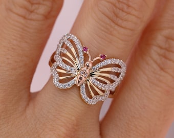 May include: A gold butterfly ring with clear and red gemstones. The butterfly's wings are detailed with intricate lines and the ring is set on a finger.