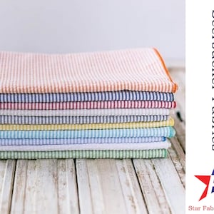 May include: A stack of colorful seersucker fabrics in various stripes. The fabrics are folded neatly and arranged on a white wooden surface. The text "Seersucker Fabrics" and "Star Fabrics Co." is visible in the image.