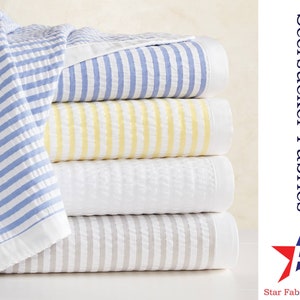 May include: A stack of four striped seersucker fabric swatches in blue, yellow, white, and gray. The fabric is soft and textured, and the stripes are a classic design. Seersucker Fabrics Star Fabrics Co.
