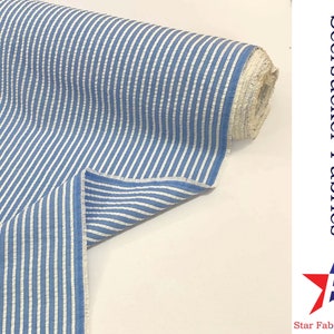 May include: Blue and white striped seersucker fabric. The fabric is folded in half and the roll is visible in the background. The text "Seersucker Fabrics" and "Star Fabrics Co." is visible in the image.