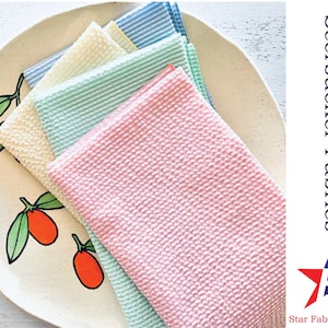 May include: Four fabric swatches in different colors, including pink, light blue, and green, with a white background. The fabric has a seersucker texture. The text "Seersucker Fabrics" is in the upper right corner of the image. The text "Star Fabrics Co." is in the lower right corner of the image.