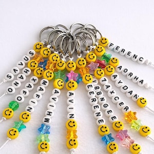 May include: A collection of metal keychains featuring white letter beads spelling names like Ferenc, Aorha, Aydan, Taxcer, Bbngee, Eerene, Avin, Avihw, Mafin, Xafo, Shano, and Savus, interspersed with yellow smiley face beads and colorful translucent star beads.