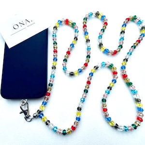 Puede incluir: Una correa de teléfono de cuentas de colores con un cierre plateado. La correa está hecha de pequeñas cuentas facetadas en tonos de rojo, verde, azul, amarillo y transparente. La correa está unida a una funda de teléfono negra con el texto "ONA. STUDIO" en la parte posterior.