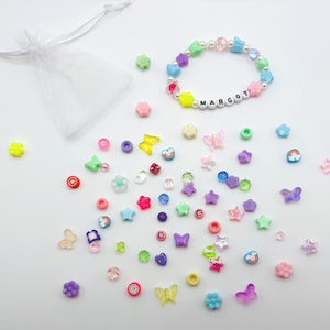 Puede incluir: Una colorida pulsera de cuentas con el nombre "MARGOT" escrito con cuentas de letras blancas. La pulsera está rodeada de varias cuentas de colores en formas de estrellas, flores y mariposas, junto con una pequeña bolsa blanca con cordón.