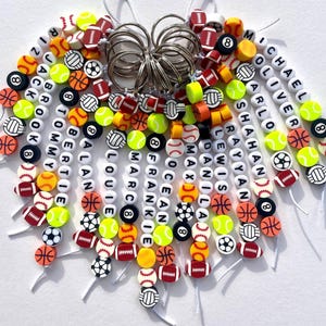 May include: A collection of beaded keychains featuring sports-themed beads and personalized name beads. The keychains include beads shaped like baseballs, basketballs, footballs, soccer balls, and tennis balls. Names like Michael, Oliver, and Jack are spelled out on the keychains.
