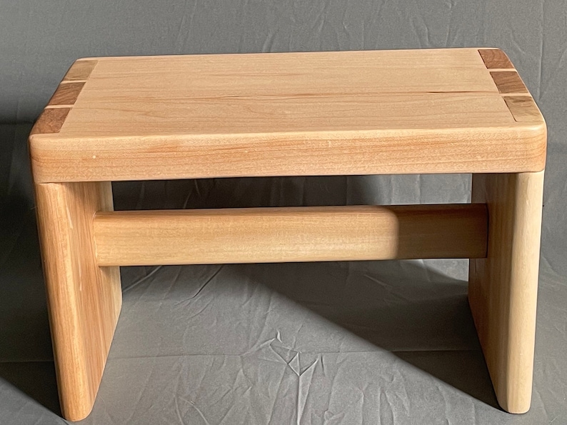May include: A small, light-colored wooden step stool. The stool has a rectangular top with rounded corners and a horizontal support beam. The legs are thick and sturdy, and the top has darker wood accents on the corners. The stool measures approximately 12 inches long, 8 inches wide, and 6 inches high.