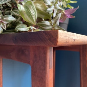 May include: A close-up of a wooden table with a rounded edge and a light brown finish. The table has a simple design with four legs and a single shelf. The table is made of solid wood and has a natural grain pattern.