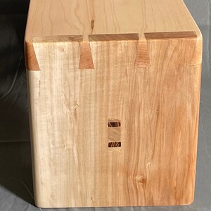 May include: A handcrafted wooden box with dovetail joints. The box is made of light-colored wood with darker wood accents at the joints. The corners are rounded, and the box has a smooth, finished appearance. A beautiful example of woodworking.