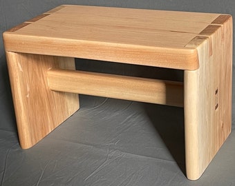 May include: A light-colored wooden bench with a rectangular top and rounded corners. The bench has a horizontal support beam and is constructed with visible joinery. The wood grain is prominent, and the bench appears to be handcrafted.