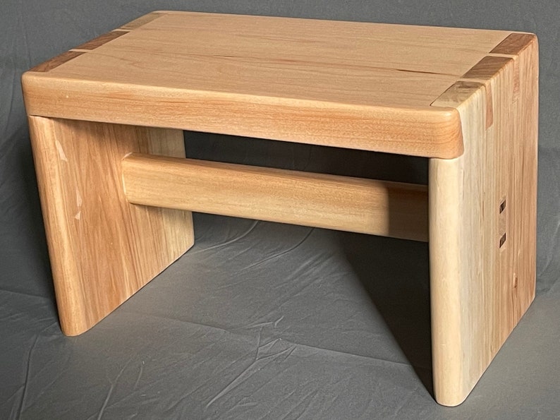 May include: A light-colored wooden bench with a rectangular top and rounded corners. The bench has a horizontal support beam and is constructed with visible joinery. The wood grain is prominent, and the bench appears to be handcrafted.