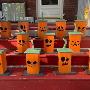 May include: A collection of 12 wooden pumpkin-shaped lanterns with different carved facial expressions. The lanterns are painted orange with green tops and are arranged on a set of red wooden steps.