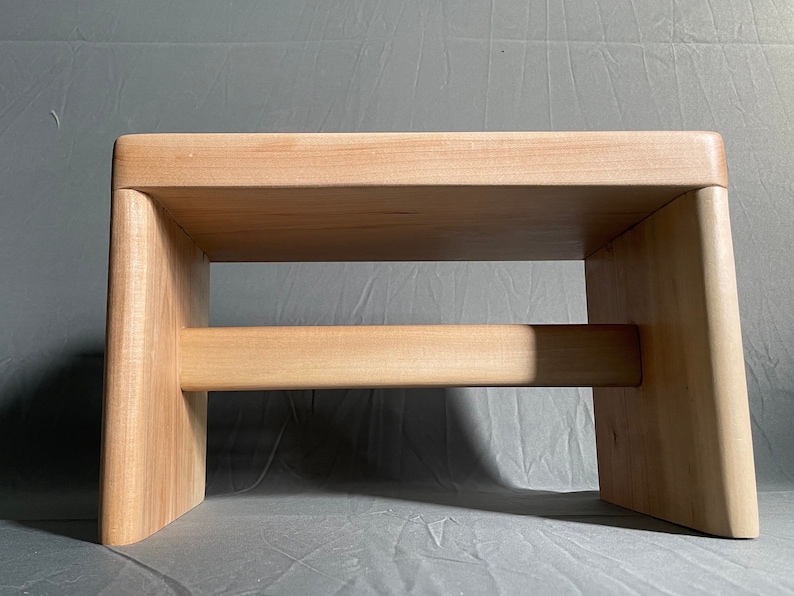 May include: A light-colored wooden step stool with a rectangular top and two supporting legs. A horizontal beam connects the legs. The stool is made of light-colored wood and has a simple, minimalist design.