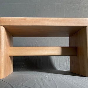 May include: A light-colored wooden step stool with a rectangular top and two vertical supports connected by a horizontal beam. The stool is made of light-colored wood and has a simple, modern design.