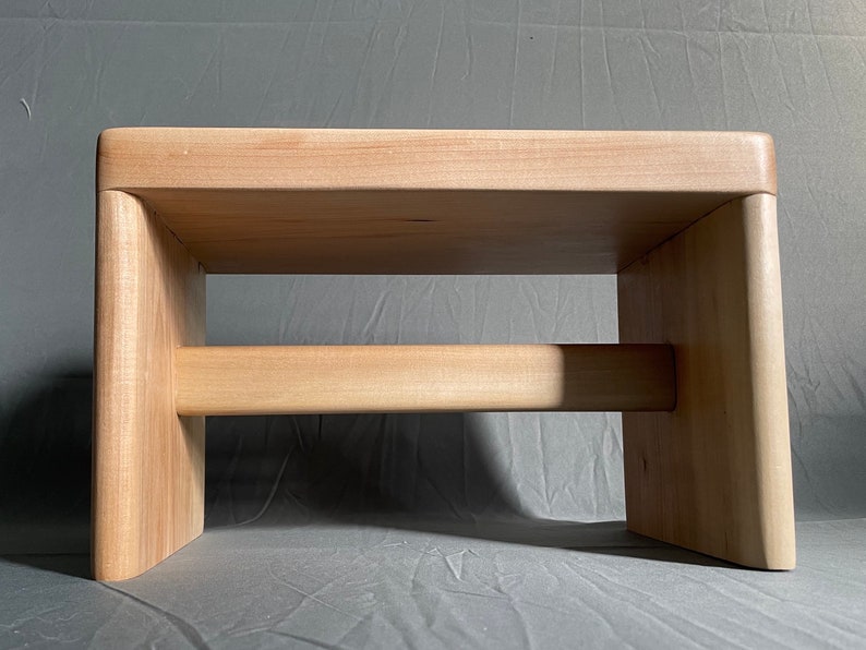 May include: A light-colored wooden step stool with a rectangular top and two vertical supports connected by a horizontal beam. The stool is made of light-colored wood and has a simple, modern design.