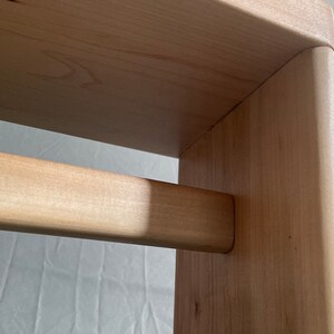 May include: Close-up of a wooden furniture piece, possibly a stool or small table. The light-colored wood has a natural grain and a smooth finish. The design features rounded corners and a horizontal support bar.