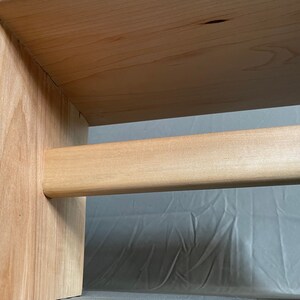 May include: Close-up of a wooden furniture piece, possibly a small table or stool. The light-colored wood has a natural grain and a smooth finish. The design features a horizontal bar connecting two vertical supports.