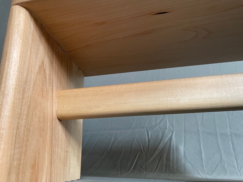 May include: Close-up of a wooden furniture piece, possibly a small table or stool. The light-colored wood has a natural grain and a smooth finish. The design features a horizontal bar connecting two vertical supports.