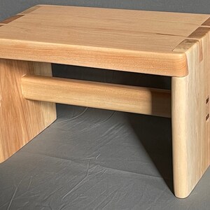 May include: A small, handcrafted wooden stool with a natural finish. The stool features a rectangular seat and a lower support beam. The joinery is visible, showcasing the craftsmanship. The stool measures approximately 12 inches long, 8 inches wide, and 10 inches high.