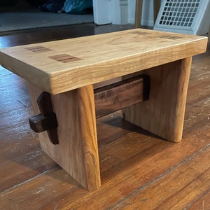 May include: A small wooden stool with a light wood top and dark wood legs. The top has a unique design with small pieces of wood inlaid into the surface.