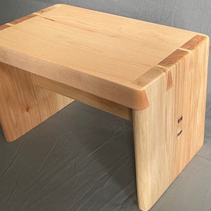 May include: A small, handcrafted wooden bench with a natural finish. The bench features a rectangular seat and angled legs, with visible joinery details. The wood has a light, warm tone, and the bench measures approximately 24 inches long, 12 inches wide, and 14 inches high.