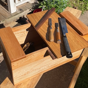 Puede incluir: Una caja de madera con una tapa con bisagras, abierta para revelar un juego de cinceles y un cepillo de mano japonés. La caja está hecha de madera clara con un acabado natural. Las herramientas están ordenadas cuidadosamente dentro de la caja.