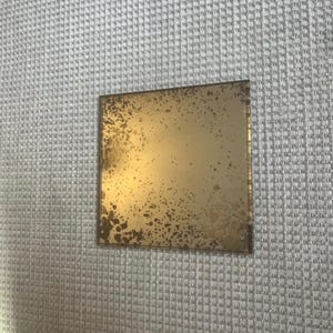 May include: Square, antique-style mirror with a gold-toned, speckled surface. The mirror has a distressed appearance, with dark spots and a slightly aged look. The edges are clean and the mirror is set against a textured, light grey background.