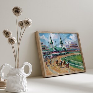 Puede incluir: Pintura enmarcada de una carrera de caballos con jinetes y caballos en la pista. La pintura tiene un marco de madera y est&aacute; sobre una superficie blanca. Un jarr&oacute;n blanco con flores secas est&aacute; en primer plano.