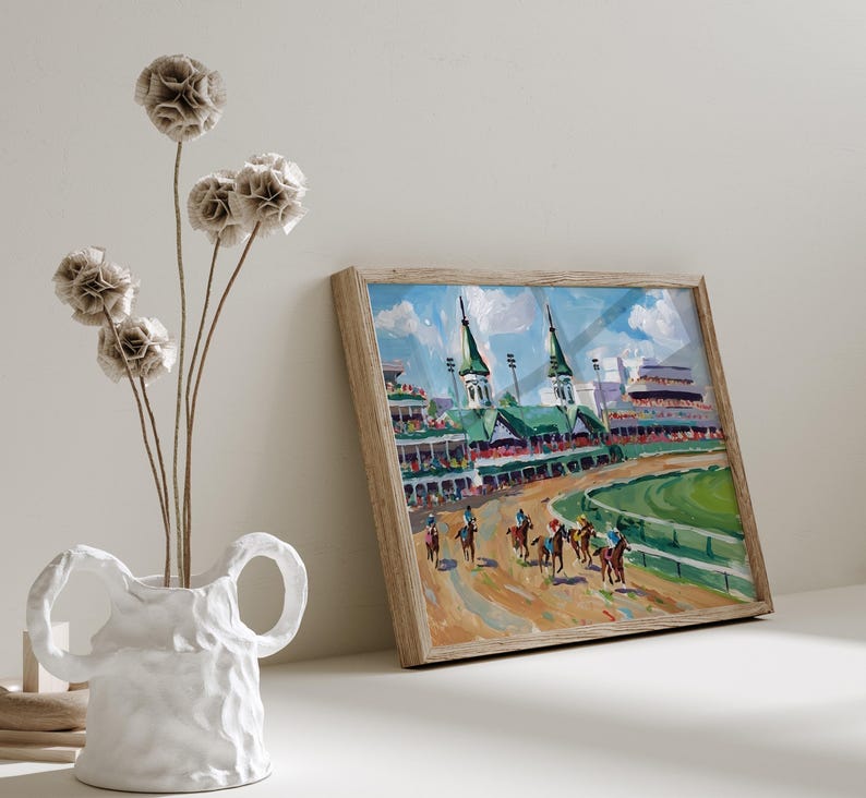 Puede incluir: Pintura enmarcada de una carrera de caballos con jinetes y caballos en la pista. La pintura tiene un marco de madera y est&aacute; sobre una superficie blanca. Un jarr&oacute;n blanco con flores secas est&aacute; en primer plano.