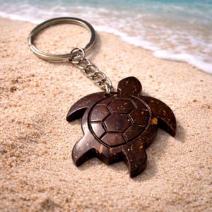 May include: A wooden turtle keyring in shades of brown, with a silver metal chain and ring. The keyring is on a sandy beach, with the sea visible in the background.