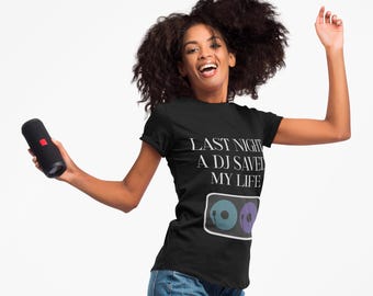 May include: A black t-shirt with the text "LAST NIGHT A DJ SAVED MY LIFE" and a graphic of two vinyl records. The person is holding a black portable speaker. The person is wearing blue jeans.