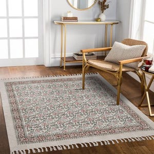 Pode incluir: Um tapete retangular com um padrão floral em tons de vermelho, verde e branco. O tapete tem uma borda creme com um design decorativo e franjas. O tapete está colocado sobre um chão de madeira.