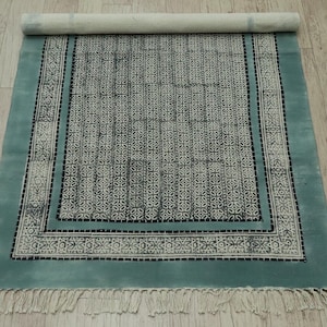 Puede incluir: Una alfombra rectangular, parcialmente enrollada, con un borde turquesa y un patrón central de motivos florales grises y blancos. La alfombra tiene flecos en el borde inferior y está sobre un suelo de madera clara.