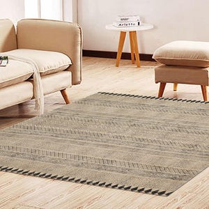 Puede incluir: Una alfombra beige y gris con un patrón geométrico y un borde de flecos. La alfombra está colocada sobre un suelo de madera en un salón.