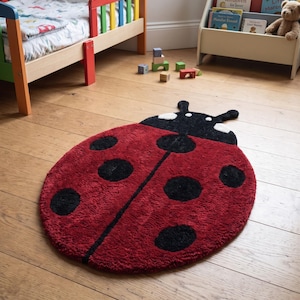 May include: A ladybug-shaped rug featuring a deep red body with black circular spots, a black head section with two white eye-like markings, and two antennae. The rug has a textured, tufted surface and is positioned on a light-colored wooden floor.