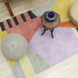 Può includere: Un tappeto geometrico colorato con un design grigio, giallo e arancione. Il tappeto è decorato con due pouf rotondi, uno grigio e uno beige, e un piccolo tavolo di legno con una ciotola blu.